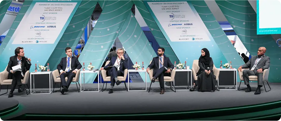 Panel discussion with five people seated on stage with teal and white banners behind them at Dubai Airshow