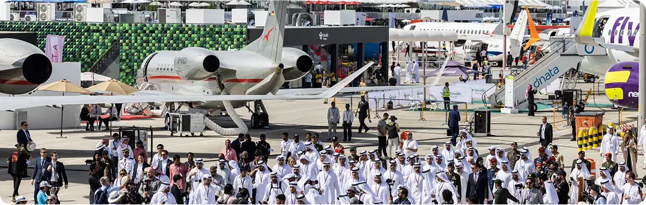 Crowd of people and jets at Dubai Airshow on tarmac with green and black patterned wall in background