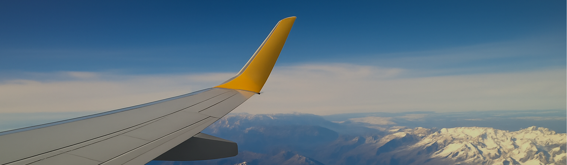 View of airplane wing with yellow tip flying over snow-covered mountains under a clear blue sky
